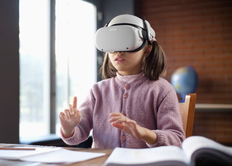 A young girl sitting at a desk and wearing VR goggles interacting with a virtual screen.